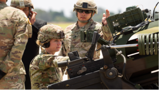 A photograph shows a female soldier and male soldiers standing beside a large gun.