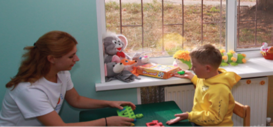 An adult and a child sit and talk at a child's table. Stuffed animals are on a shelf between them.