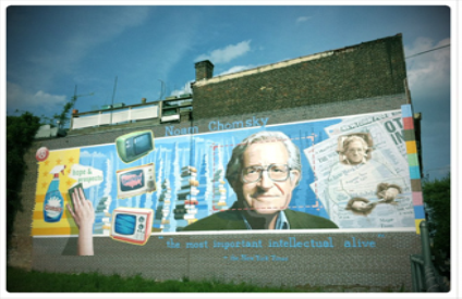 A photograph shows a mural on the side of a building. The mural includes Chomsky's face, along with some newspapers, televisions, and cleaning products. At the top of the mural, it reads “Noam Chomsky.” At the bottom of the mural, it reads “the most important intellectual alive.”